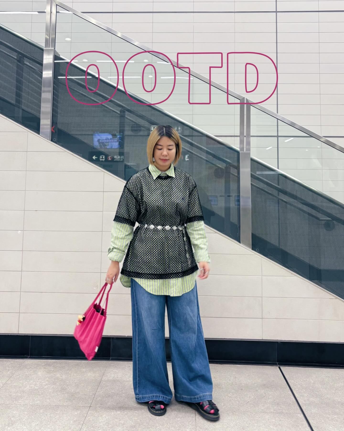 The image shows a person standing in what appears to be a shopping mall or commercial building with white tiled walls and an escalator visible in the background. They're wearing a layered outfit consisting of a light green striped shirt underneath a black mesh overlay top, wide-leg blue jeans, and dark shoes. They're accessorized with a bright pink handbag and what looks like a decorative belt or waist detail. Behind them, part of a sign with "OOTD" (outfit of the day) is visible in pink lettering against the white wall.