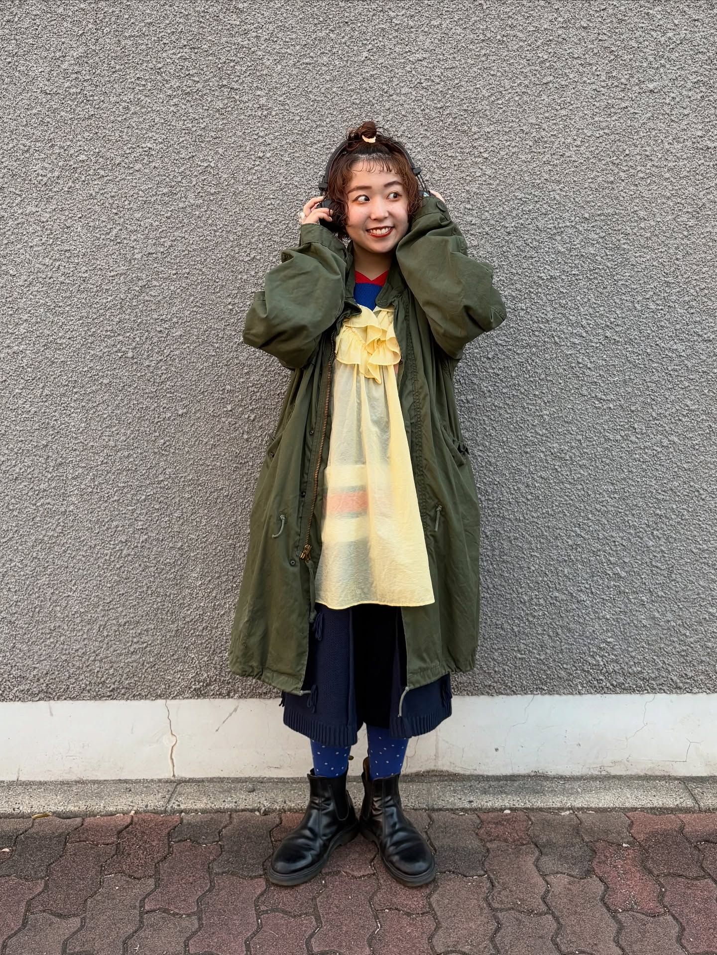 The image shows a person standing against a gray wall, wearing a long olive green coat over a yellow dress with some decorative elements. They're also wearing blue jeans and black boots. The person has their hands raised near their head in a playful pose. They're standing on a brick or paver stone surface in what appears to be an outdoor setting.