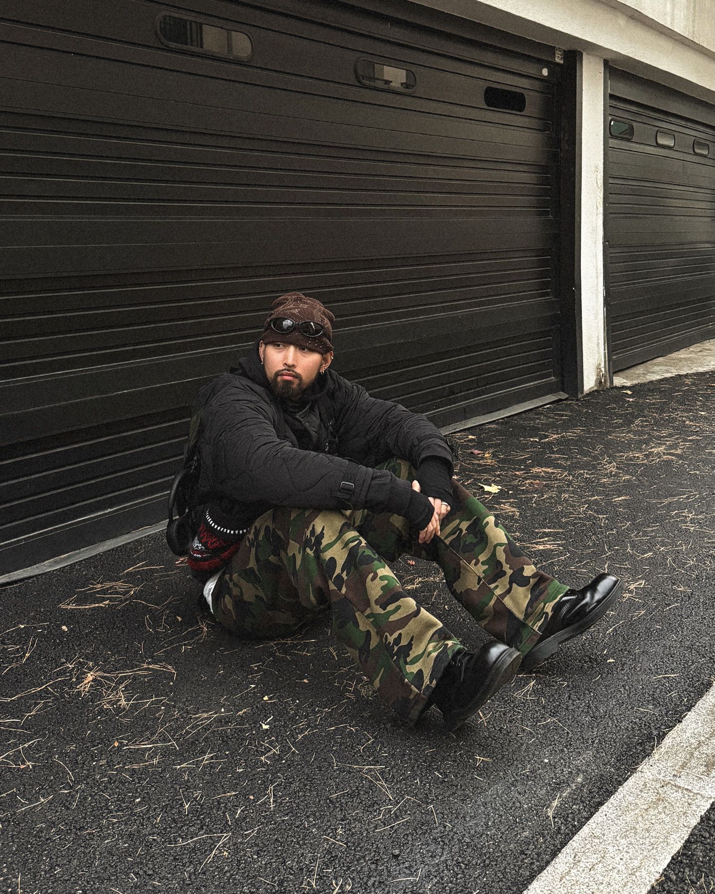 A person sitting on the ground in front of black garage doors. They are wearing camouflage pants, a black jacket, and a dark cap. The person is seated on asphalt with some fallen leaves scattered around.