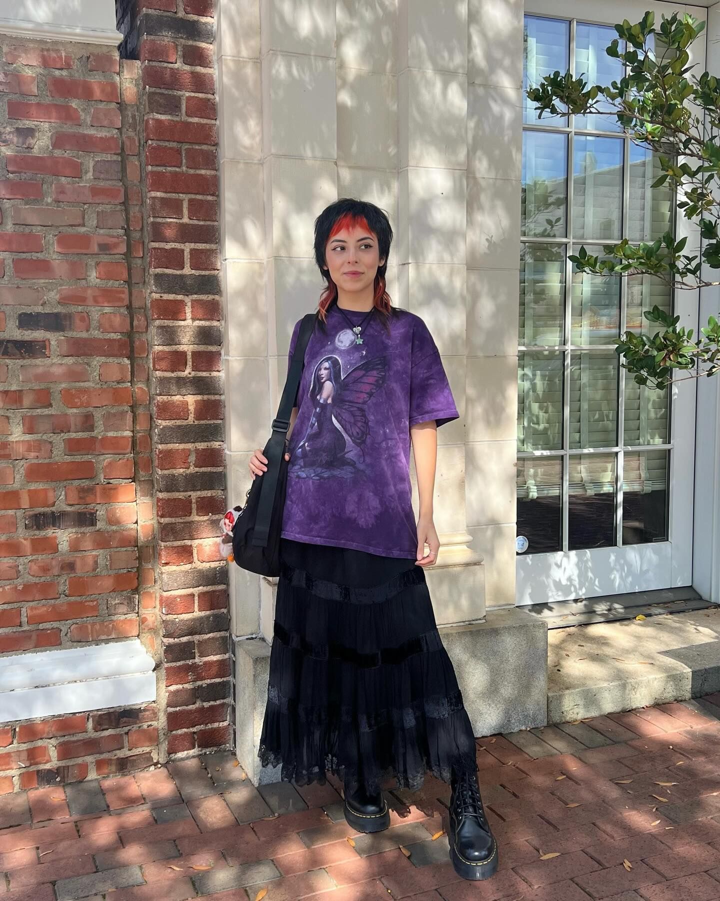 A person standing outside a building with brick walls and white trim. They're wearing a purple graphic t-shirt paired with a flowing black tiered maxi skirt and black boots. They have short dark hair with reddish highlights and are carrying a black shoulder bag. Dappled sunlight creates shadows across the scene, with green foliage visible near a large window.