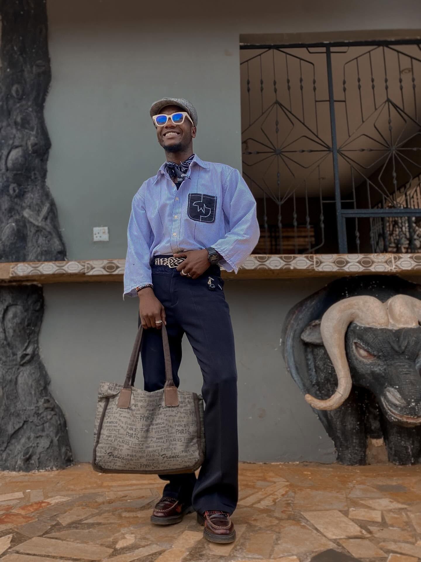A person standing outdoors in front of a building with decorative gates. They're wearing sunglasses, a light blue button-up shirt with a pocket, dark pants, and brown shoes. They're holding a woven tote bag and smiling. The setting appears to be a stone-paved area with decorative stone elements visible on either side of the frame.