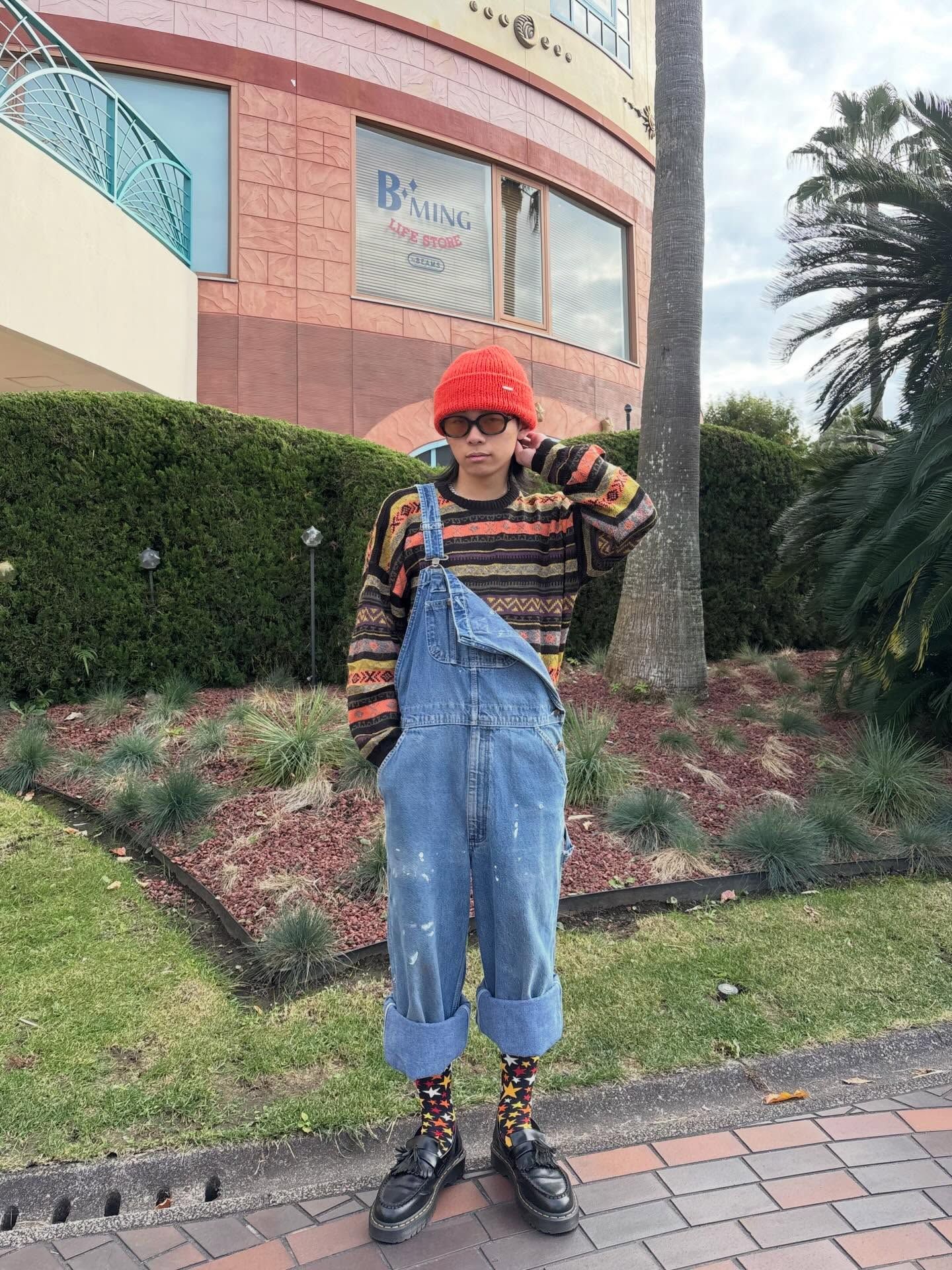 The image shows a person standing on a brick pathway in front of a commercial building with pink walls. They're wearing an oversized denim overall with rolled-up cuffs, a colorful striped knit sweater, patterned socks, and black boots. They have on a bright red knit beanie/watch cap and sunglasses. Behind them is landscaping with shrubs, palm trees, and a grassy area. The person is posing with one hand adjusting their hat.