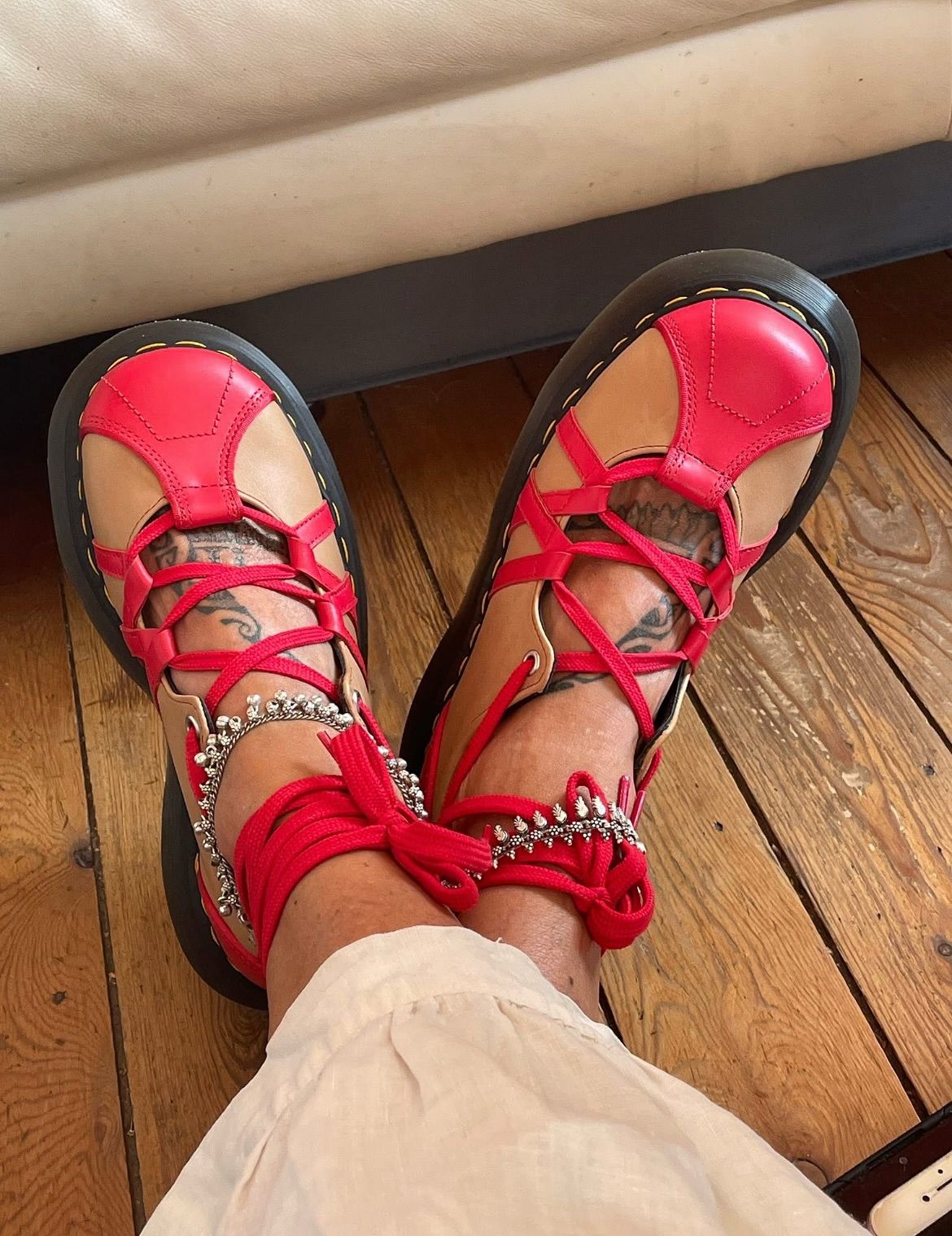 A pair of bright red Dr. Martens shoes with a distinctive design featuring cutouts and straps. The shoes have black soles and are being worn by someone in light-colored pants. The footwear is displayed on a wooden floor, with what appears to be a light-colored furniture piece visible in the background.
