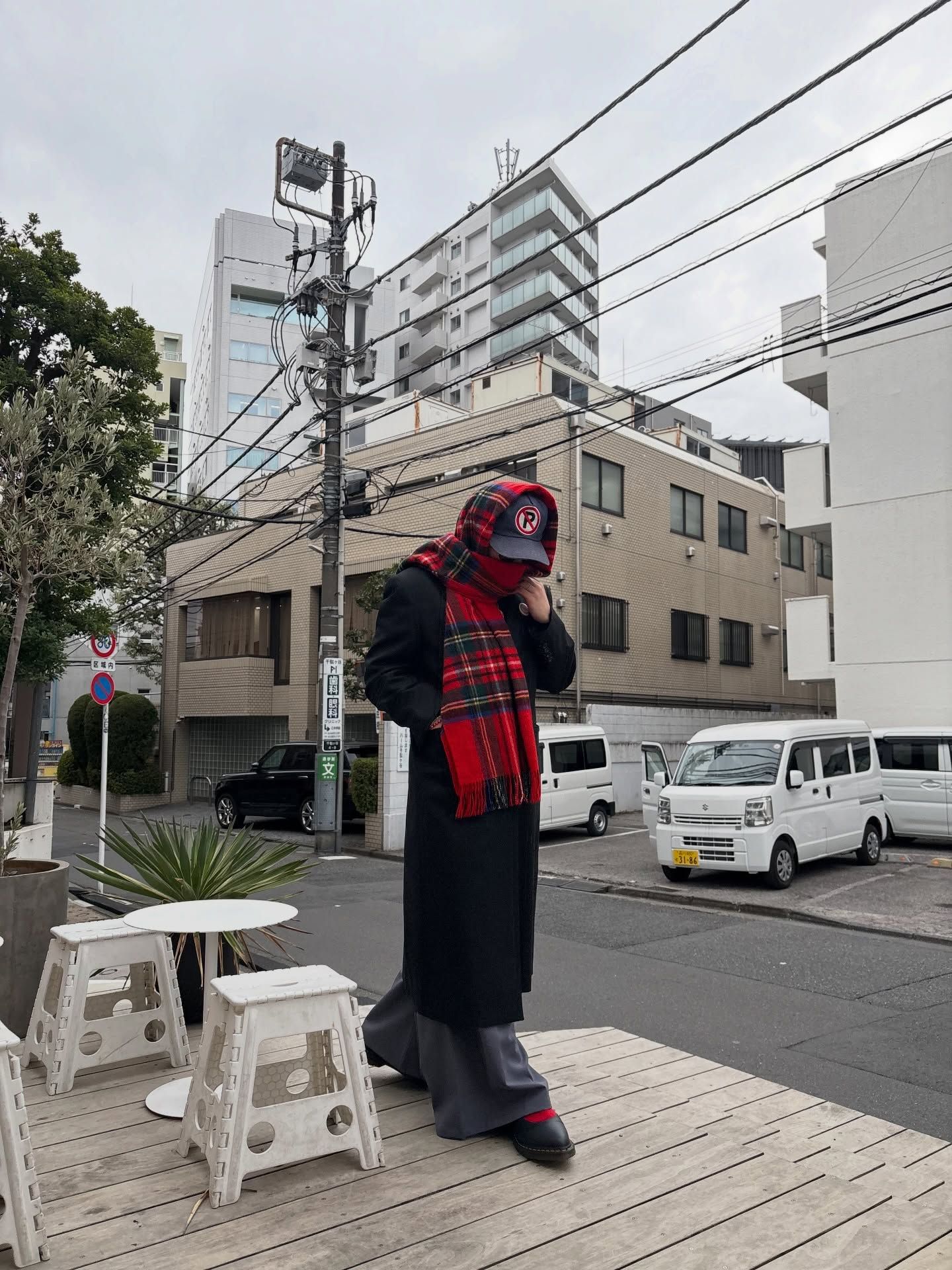 A person standing on a wooden deck in an urban Japanese setting, wearing a long black coat, bright red and black plaid scarf, and matching cap. They're positioned near white stools with apartment buildings and utility poles visible in the background. Several parked vehicles including white vans can be seen on the street. The scene appears to be on an overcast day in a residential neighborhood.
