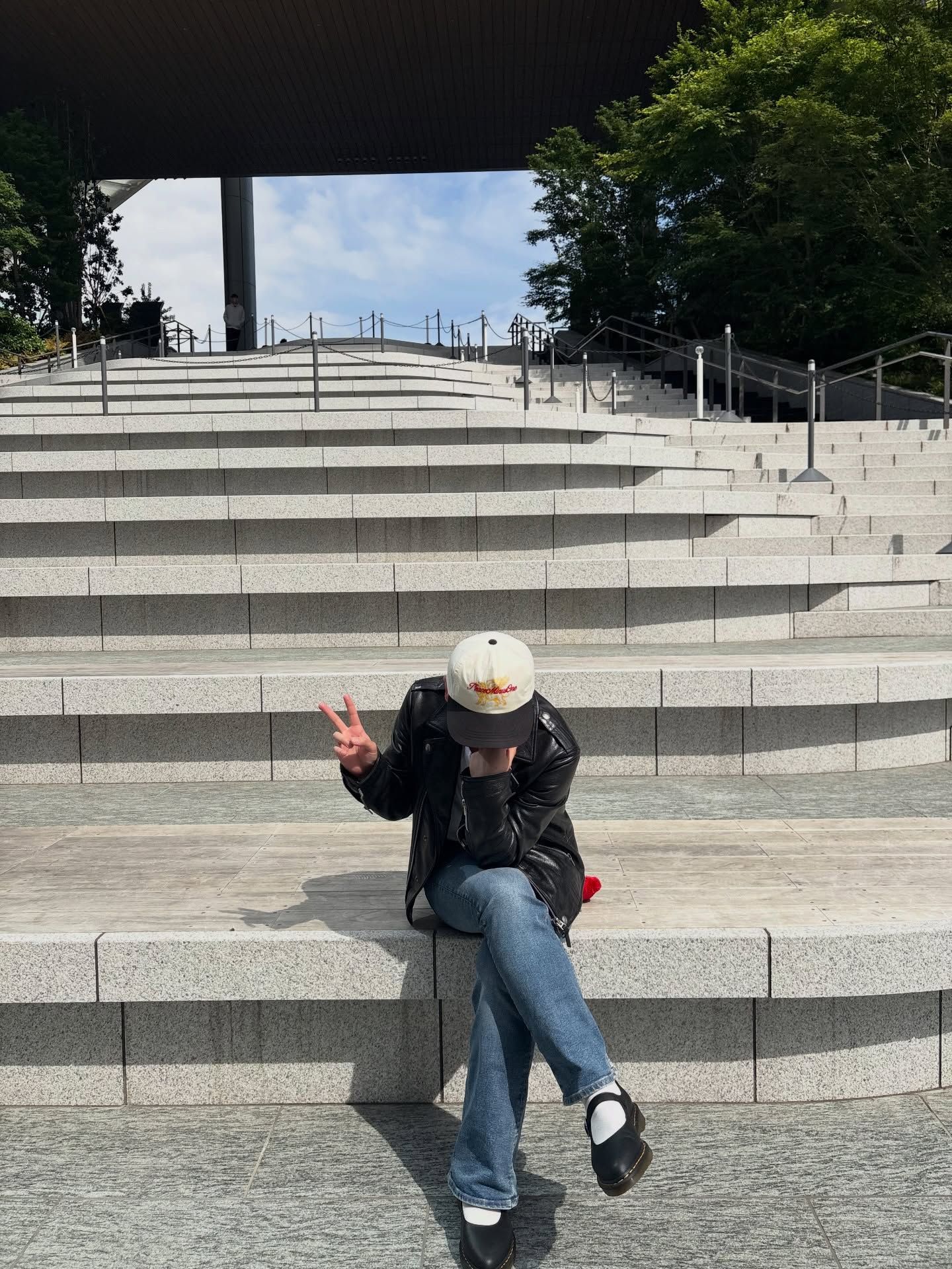 A person sitting on wide outdoor stone steps, wearing a black leather jacket, blue wide-leg jeans, black platform shoes, and a cream-colored baseball cap. They are making a peace sign gesture with their hand while holding a red object. Lush green trees and a blue sky are visible in the background.