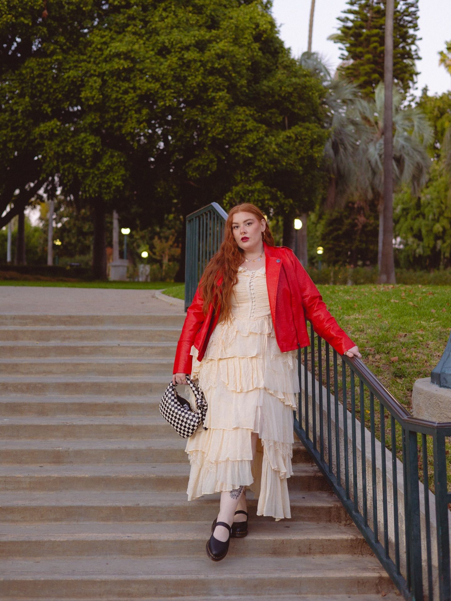 A person with long red hair descending stone steps in a park setting. They are wearing a layered cream-colored ruffled dress with a bright red leather jacket and dark shoes. They're carrying a checkered handbag and holding onto a metal railing. The background shows green trees and landscaping at dusk with some lights visible.